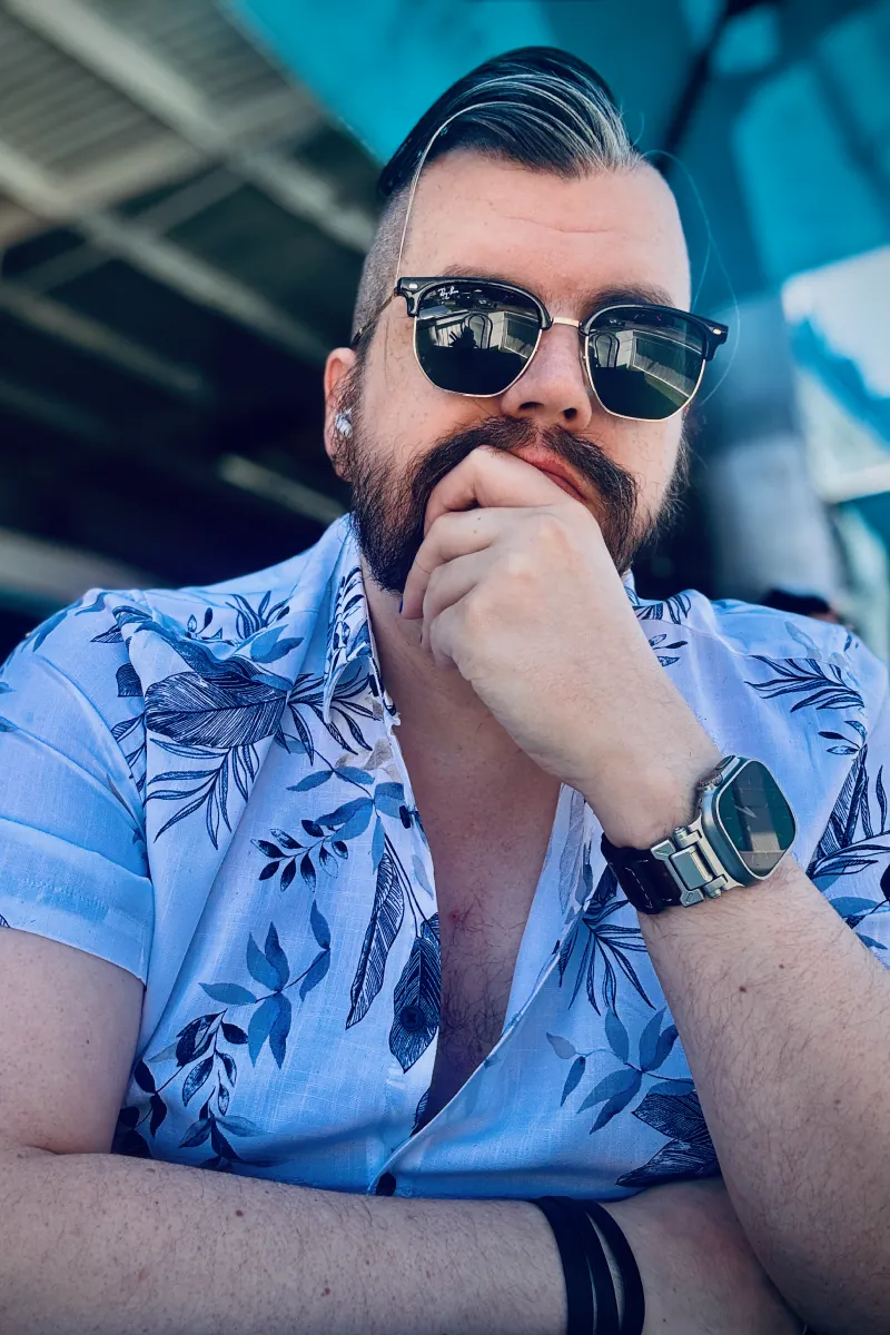 Portrait of me, Jono. I'm sitting at a table in a white shirt with a floral pattern, resting my chin on my left hand. I've got a beard and slicked-back brown hair with a grey streak on the front-left. I'm wearing Ray-Ban clubmaster style sunglasses, an Apple Watch Ultra with a metal and leather band on one wrist, and a thin leather strap wrapped around the other wrist.